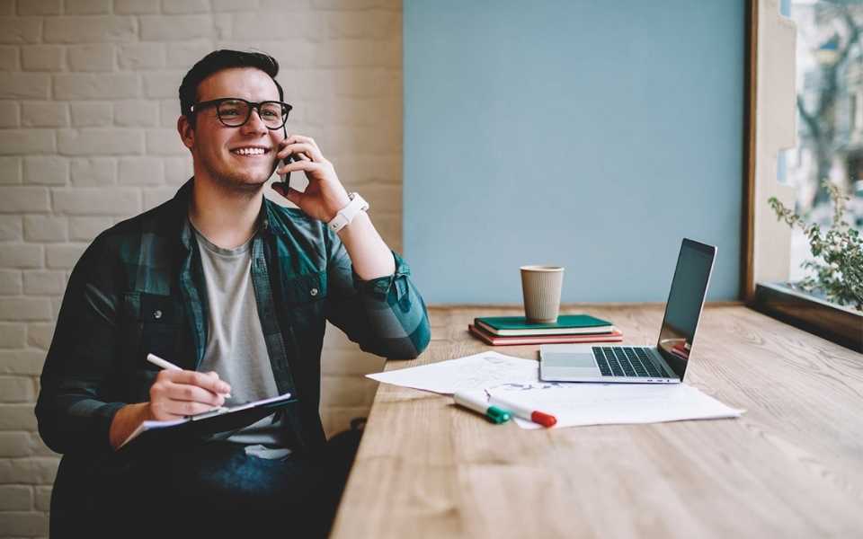 An employee wearing glasses talks on the phone while typing on laptop. Concept of freelance translators.