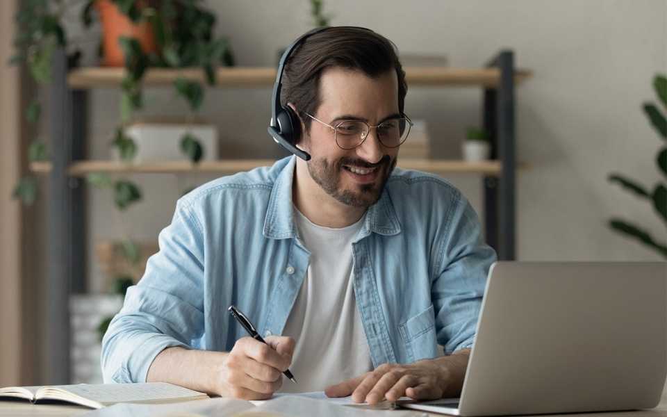 A man with headphones smiling, taking notes and using laptop. Concept of language skills and development.
