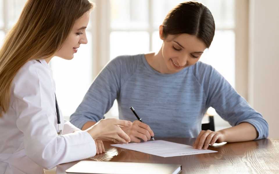 A woman and a doctor sitting with the woman holding the pen and writing. 