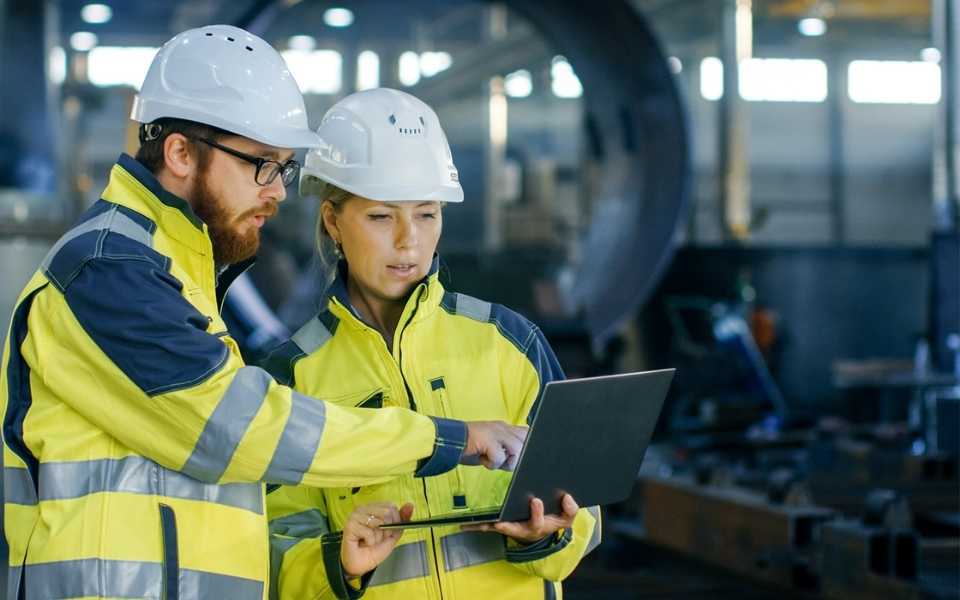 Two engineers wearing protective helmets, and one is carrying a laptop. Concept of engineering translations.