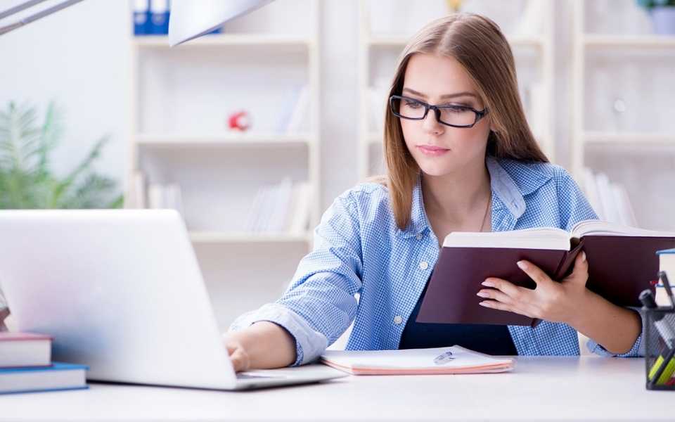 A girl wearing glasses and holding a book while using a laptop. Concept of online editing and proofreading services.