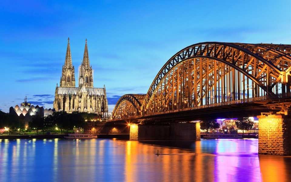 Night-time skyline of Cologne, Germany, with an iron bridge over a river. Concept of German translation services.