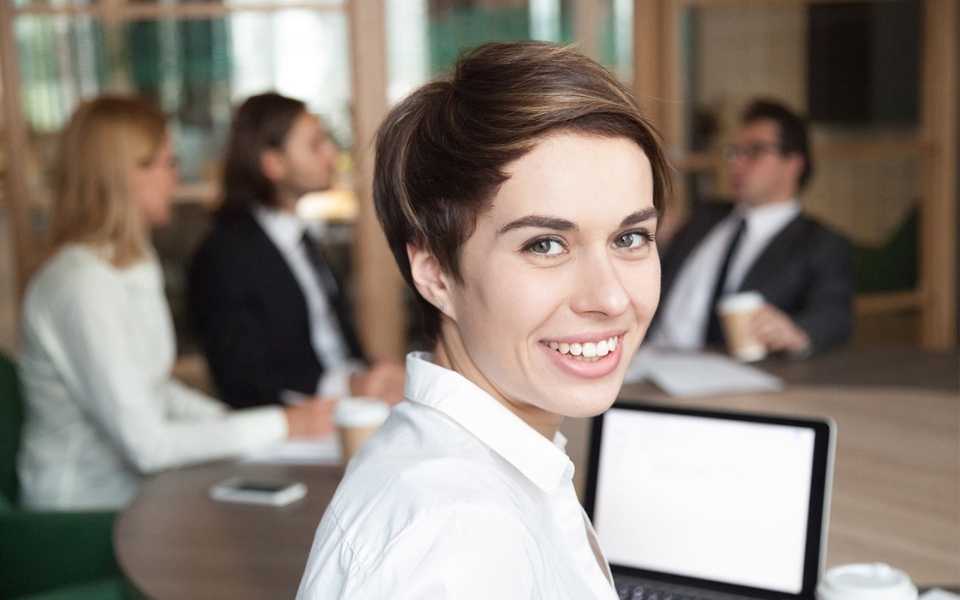 Brown-haired girl smiling with co-workers behind her at a business meeting. Concept of language translation services office.