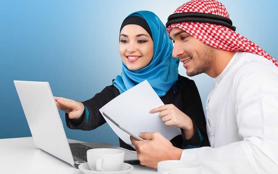 A girl wearing hijab and a man wearing traditional Arabic clothing, sitting on a desk with a laptop. Concept of English to Arabic translation.