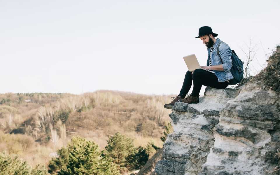 A man wearing a backpack sits on a cliff using his laptop. Concept of freelance translators working for translation agencies.