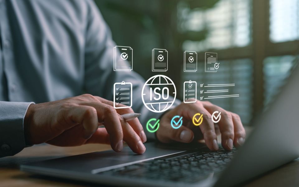 A businessman is using a laptop to illustrate quality control and international standards with ISO certification icons, a checklist, and compliance paperwork. ISO-certified AI language technology. 