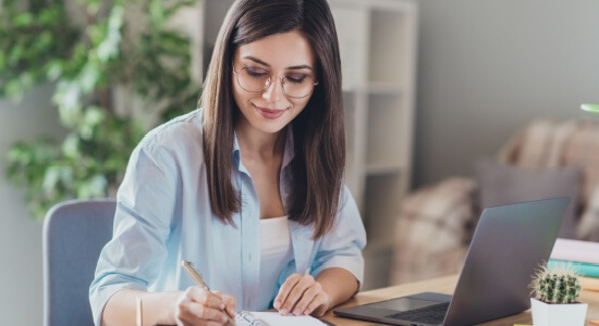 A lovely young girl wearing spectacles is using a laptop, taking notes, and holding a pen. A concept of professional Albanian translators.
