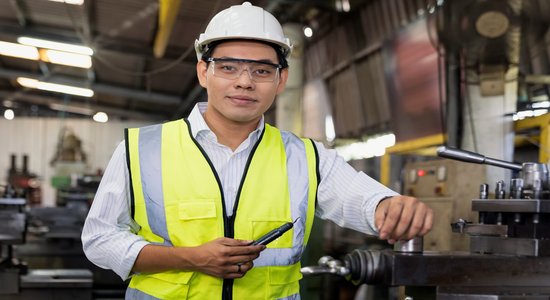 Man in white hard hat with a tool leaning against workshop machinery.
