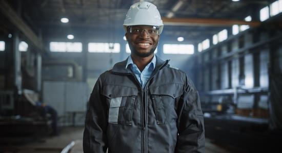 A dark-skinned engineer in a hard hat and goggle smiling at camera. 