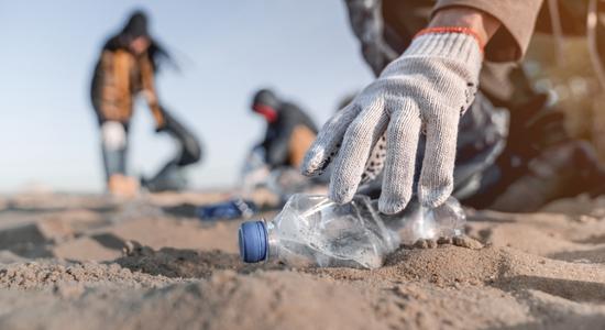 A gloved hand picking up a crushed plastic bottle on sand.