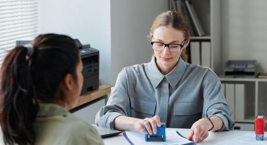 A woman stamping paperwork as another observes. 