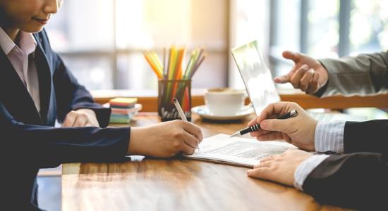 Three people pointing at and signing a contract at a desk. 