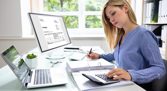 Blonde woman working on desk using computers and calculator.