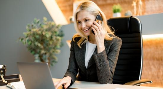 A blonde businesswoman smiling with a phone and a laptop. 