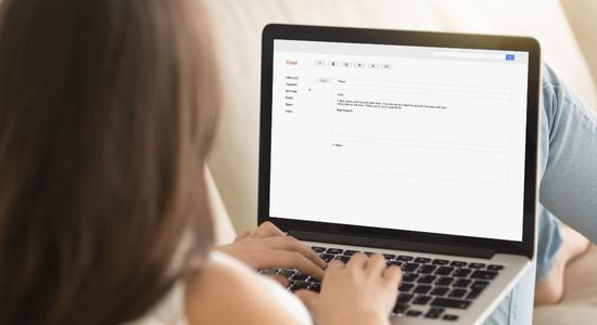A woman reclining on a couch, typing an email on her laptop. 