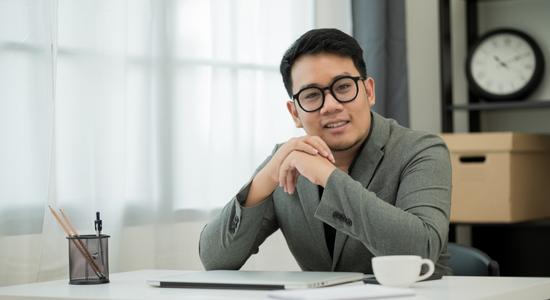 A bespectacled man with folded hands at a desk smiling at camera.