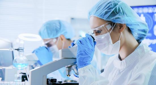 Two scientists in lab gear looking in white microscopes.