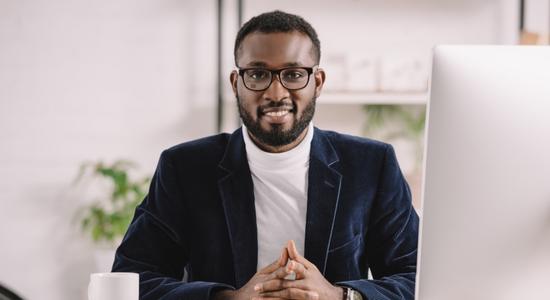 A dark-skinned, bespectacled man at a desk, smiling at camera. 