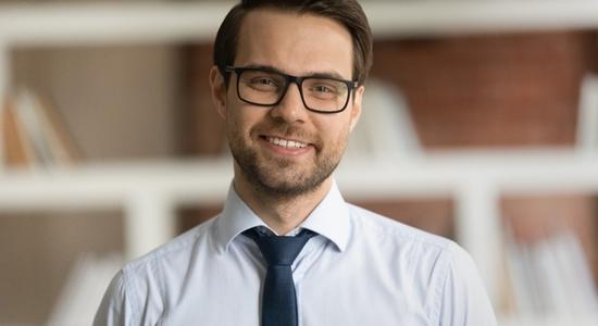 A bespectacled man in dress shirt and tie smiling at camera. 