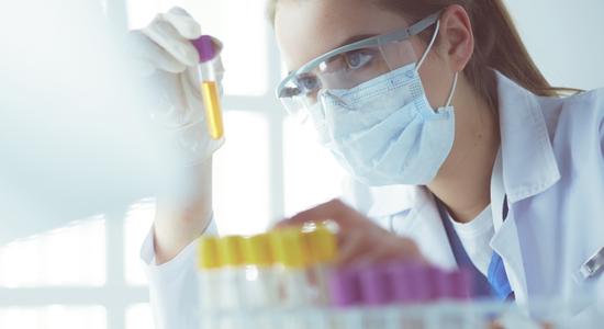 A woman in lab gear closely inspecting test tubes containing yellow fluid.