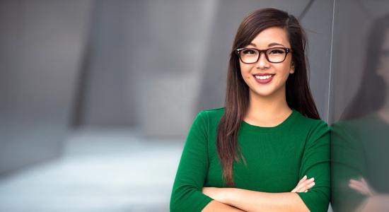 A bespectacled woman in green leaning against a wall, smiling at camera.