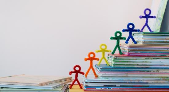 Rainbow coloured felt stickmen on ascending stacks of books.