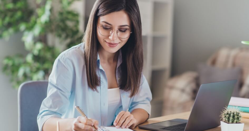 Beautiful young girl in a shirt sitting at a desktop computer, holding a pen and writing on a notepad and wearing glasses. A concept of document translation services.